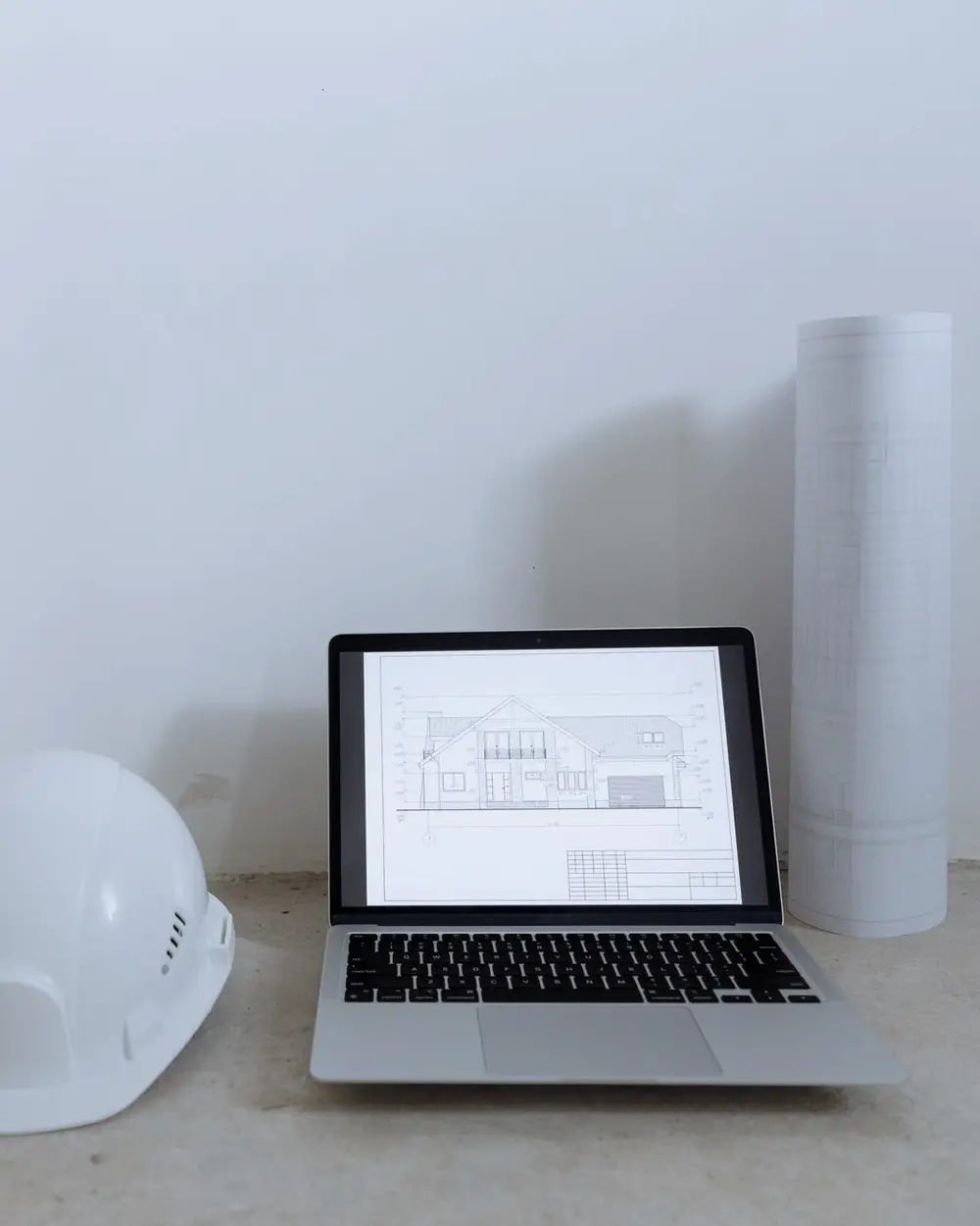 Notebook con un plano en pantalla, un casco y planos enrollados sobre el piso de hormigón de una obra